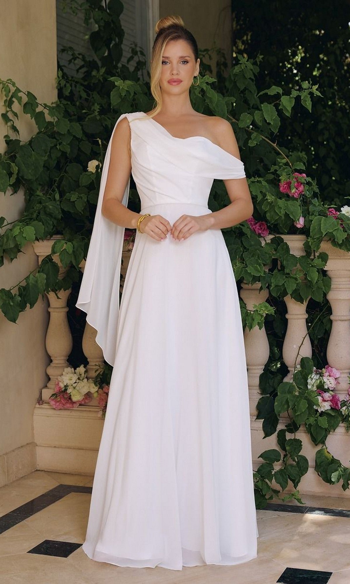 A woman stands in a leafy garden wearing the Asymmetrical Off-White Bridal Gown: Amoris T716W, featuring a long chiffon skirt, draped fabric, and minimal accessories.